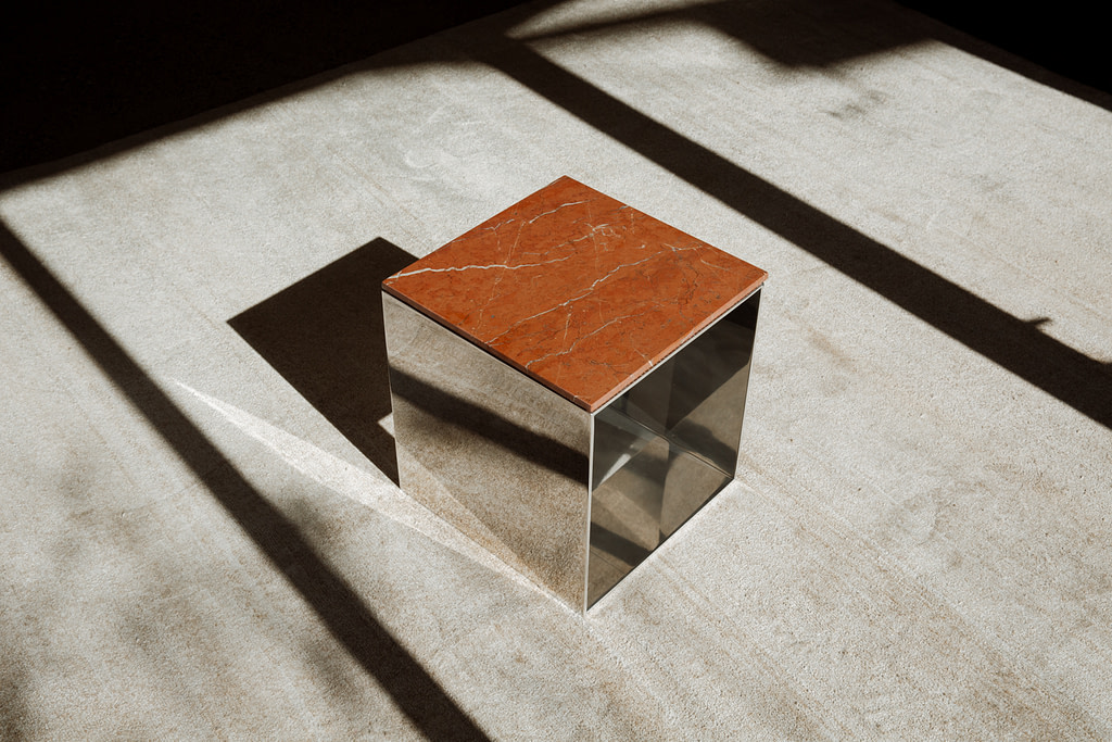 Material Studies Cube v.1, mirrored aluminum, Iron Red stone top, low three-quarter, hard shadows across the floor