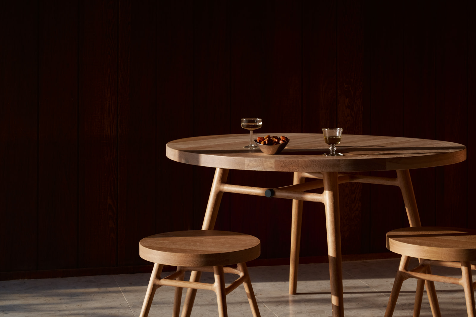 Bough Table Round and two Bough Stools in white oak in a dark wood-paneled interior, coupe glasses and a bowl of nuts on top.