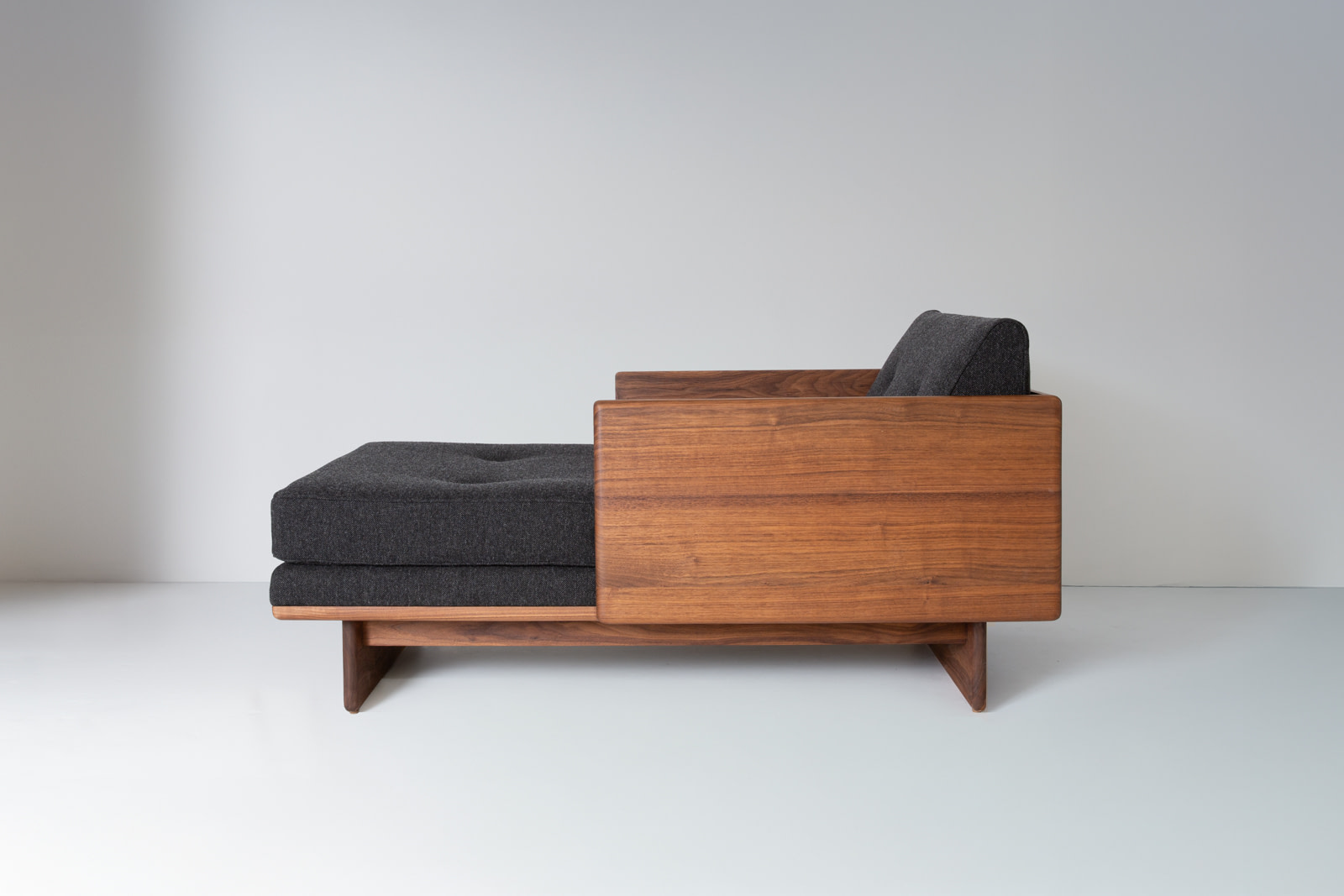 Kalonstudios no6 chaise walnut studio side Mid century Wooden Sofa with Dark Gray Cushions Against a Light Wall Shown from the Side