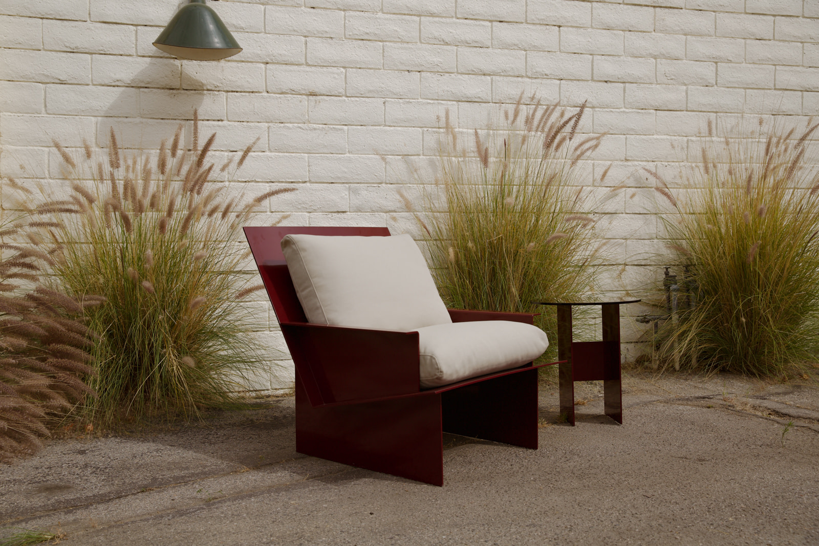 Maroon metal outdoor chair with cream cushions and a small matching table, set against a white brick wall with tall ornamental grasses.