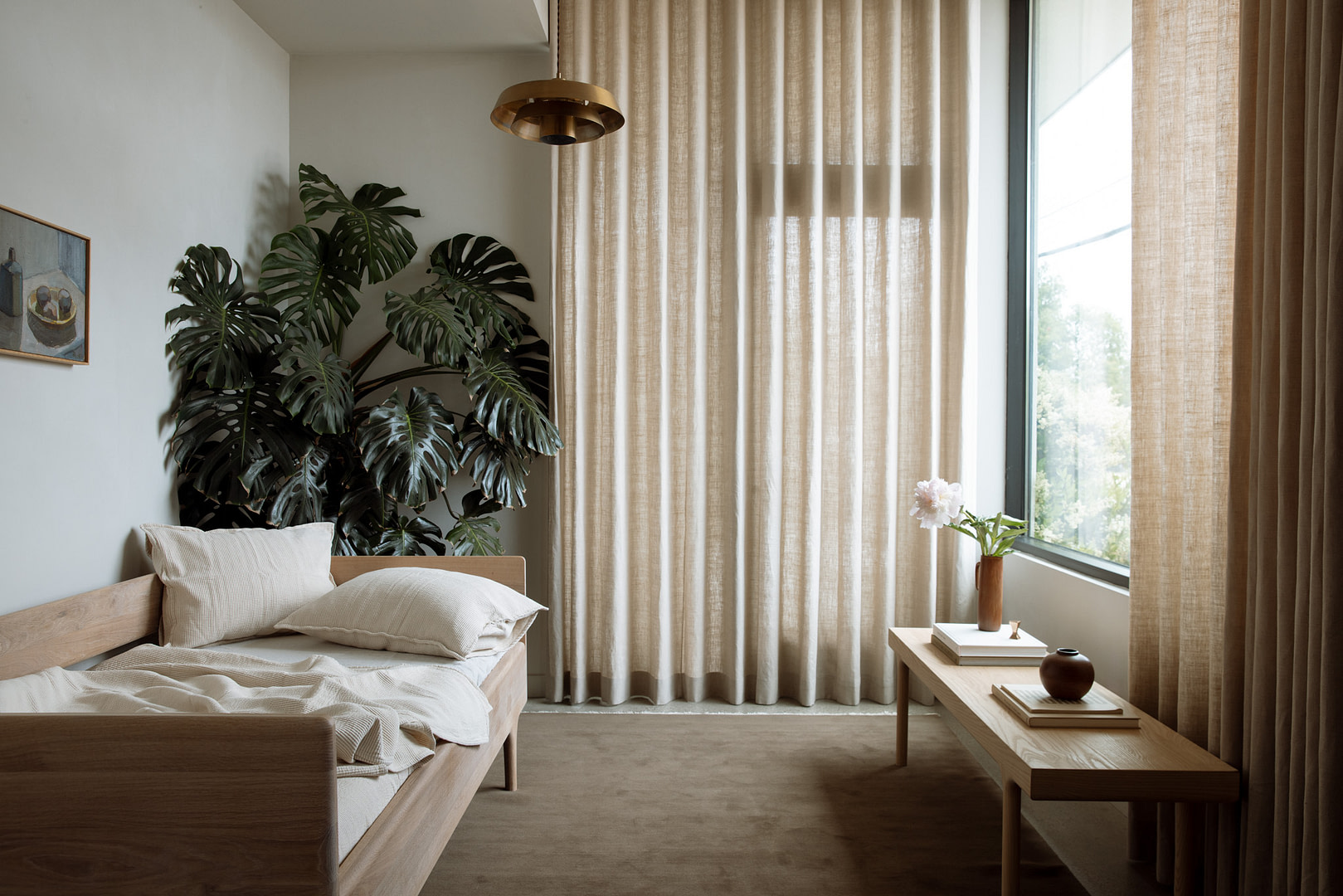 Sunlit Bedroom Featuring a Wooden Bed with Beige Linens a Large Monstera Plant and Light Beige Curtains by a Window |  | Kalon Studios Us