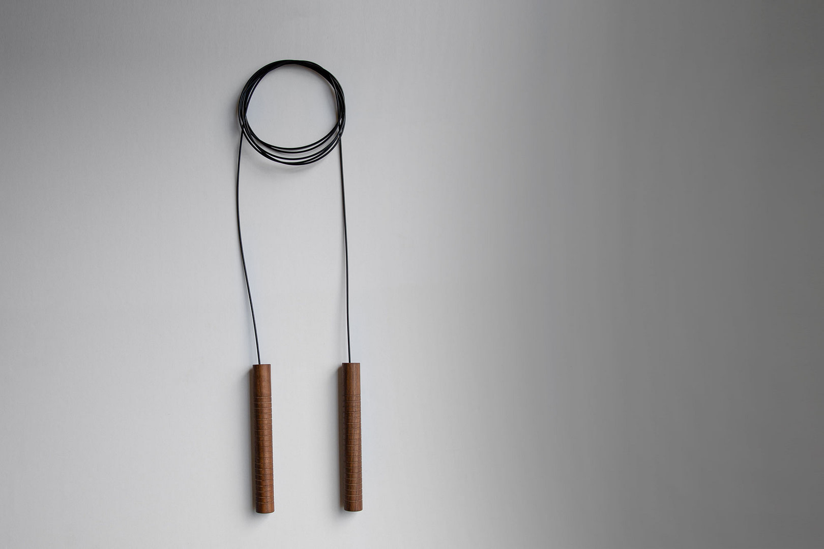 Jump Rope Jump Rope in Walnut with Grooved Handles and a Black Cord Coiled at the Top Hanging Against a Gray Wall