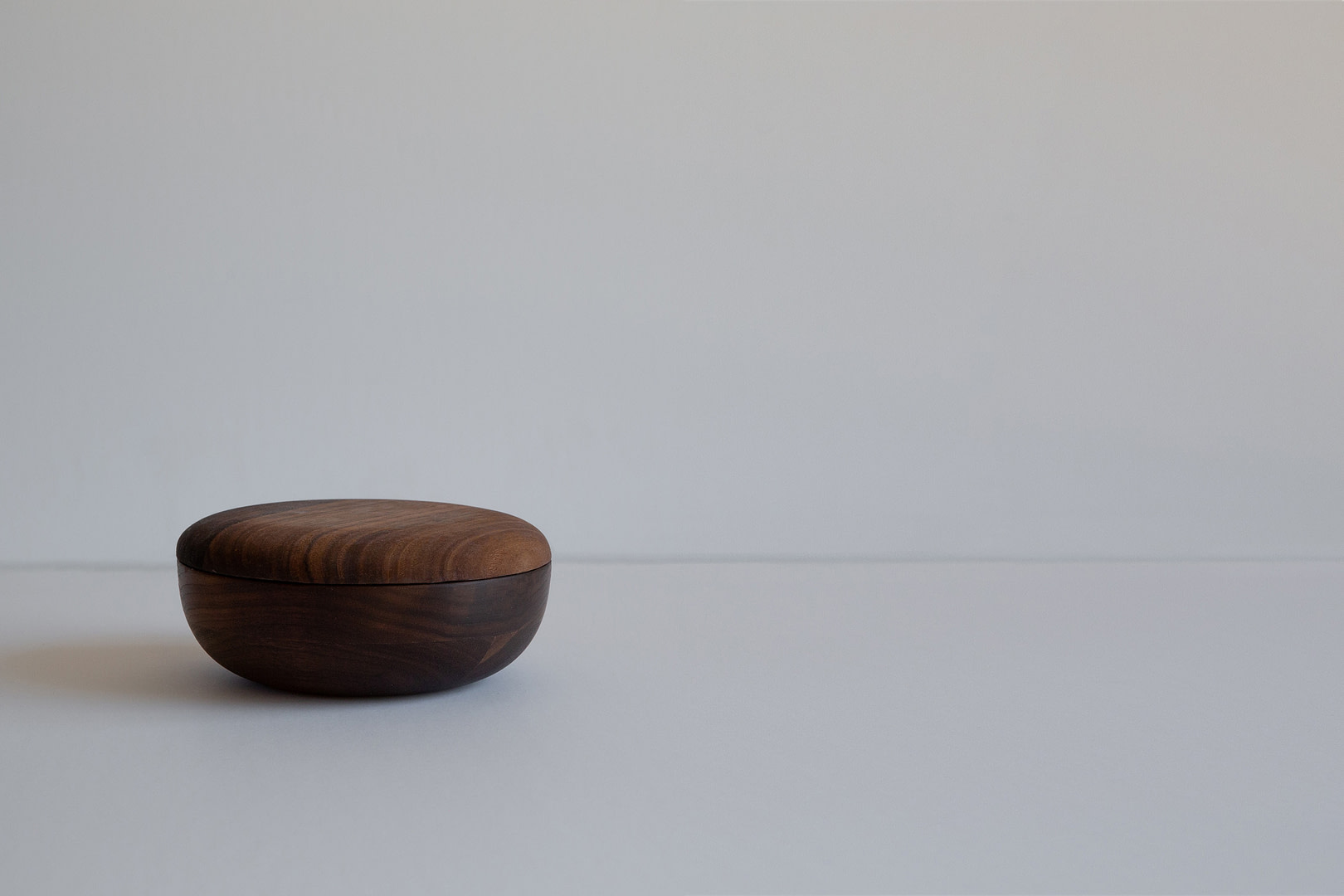 Kalon woodentableware walnut 6inchbowl 6 inch Roasted Bowl with Lid On a Small Dark Rounded Form Set to One Side of a White Field