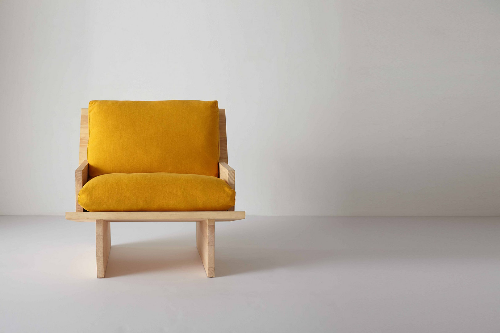 Rugosa Chair in sugar pine with golden-yellow linen cushions, set left on a white studio floor, slab arms and panel-leg base.