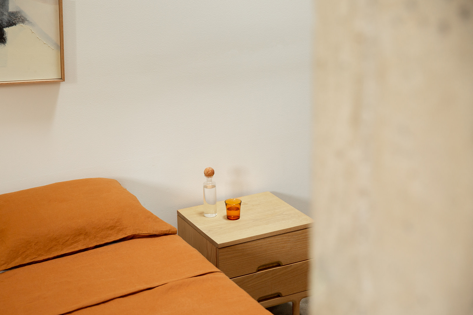 Bedroom with orange bedding, a wooden nightstand, a corked bottle and amber glass, beige walls, and a sheer curtain.