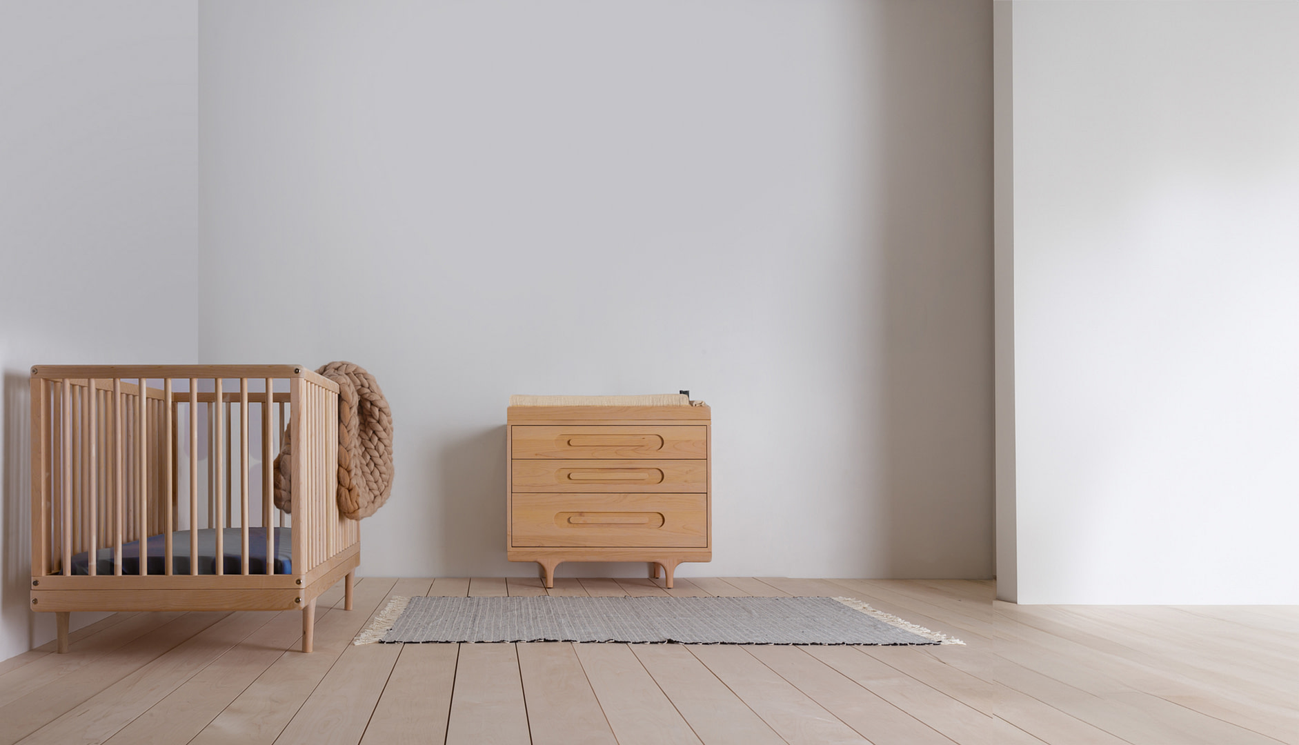 Kalonstudios caravan bundle whiteoak naturalfinish leftaligned Wooden Baby Crib on the Left with a Chunky Knit Blanket Draped over Its Side in a Pale Minimalist Nursery