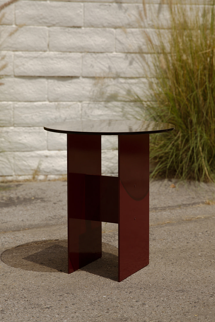 Round glass-top table with a burgundy metal base outdoors against a white brick wall and grasses in the background.