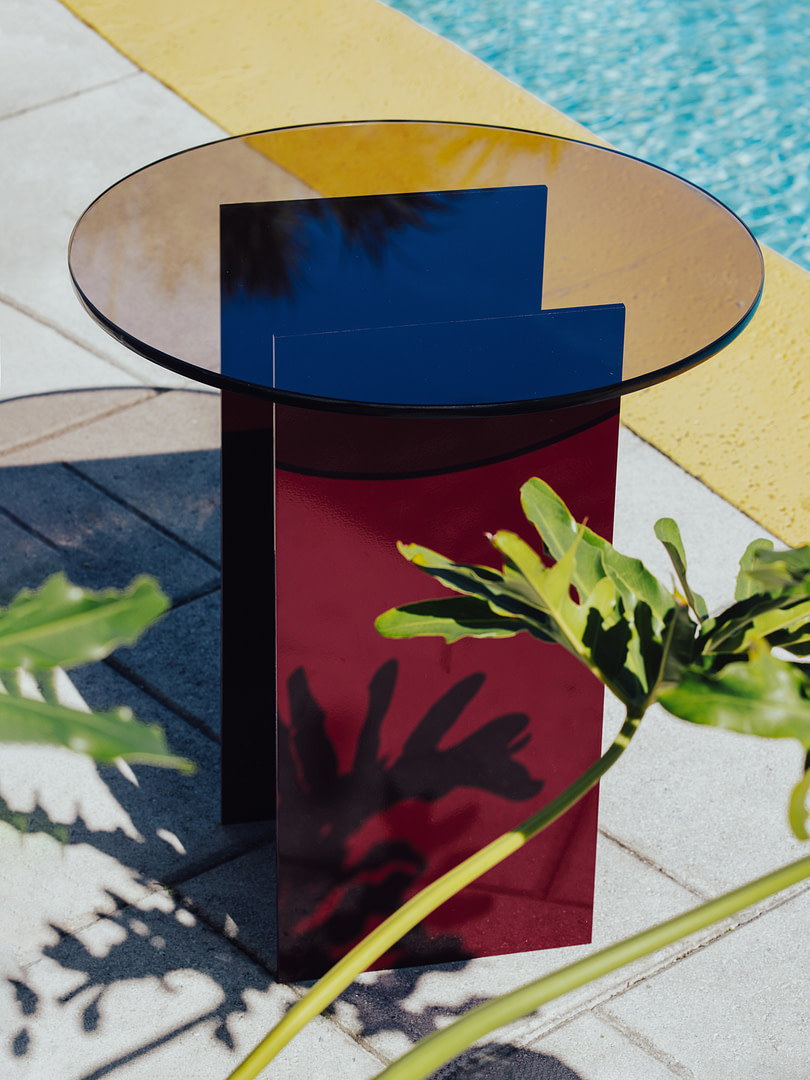 Modern outdoor sculpture with red glossy pedestal, blue geometric panels, and a round glass top beside a poolside setting.