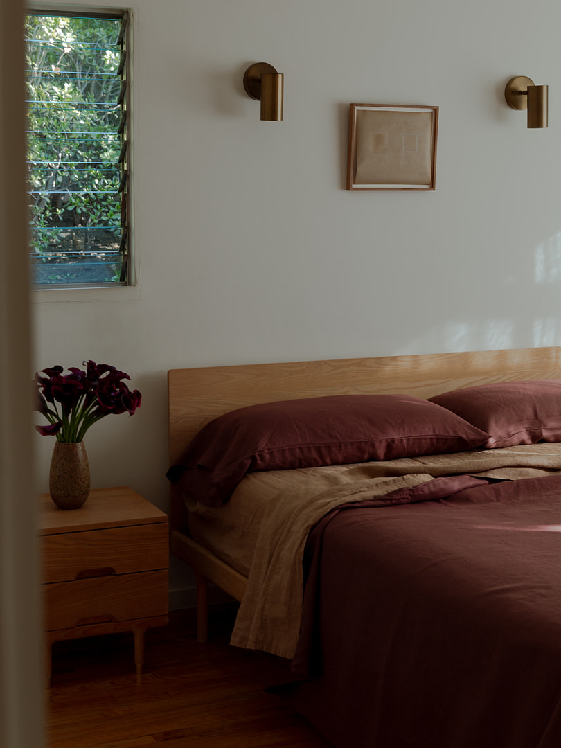 Wooden bed with burgundy bedding and a matching nightstand, a vase of purple flowers on the side, and a window with slatted blinds to the left.