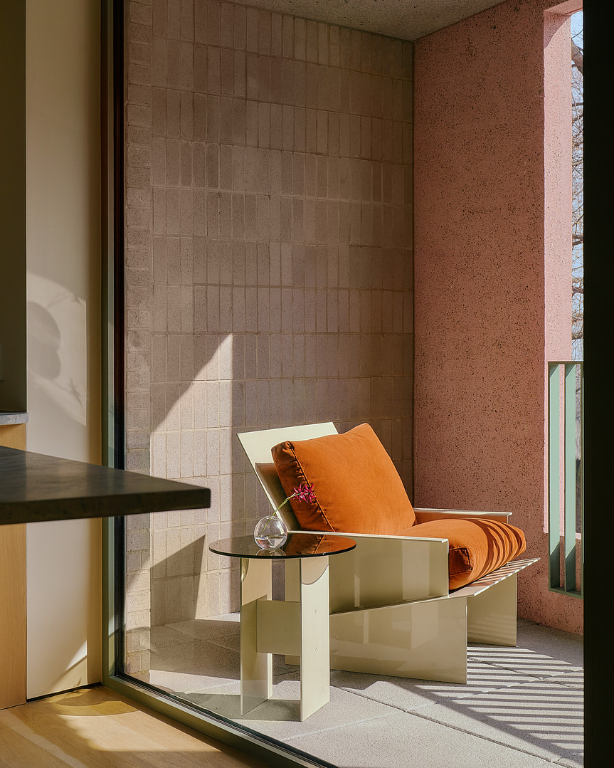 Kalon Material Studies I Aluminum Outdoor Furniture 4 Outdoor Balcony Seating with an Orange cushioned Chair Glass Round Table and Small Vase with a Pink Flower Against a Brick Wall