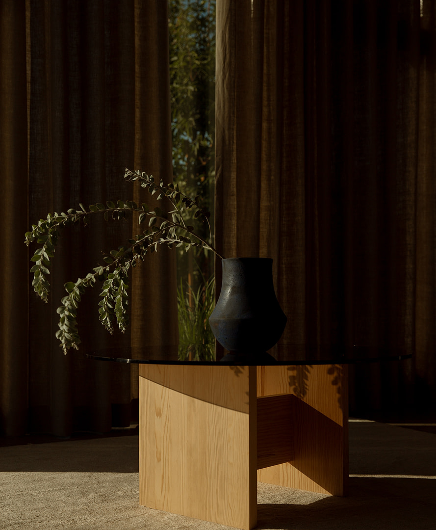 Rugosa Coffee Table in sugar pine with a bronzed-glass top, dark ceramic vase and a trailing branch, linen curtains behind