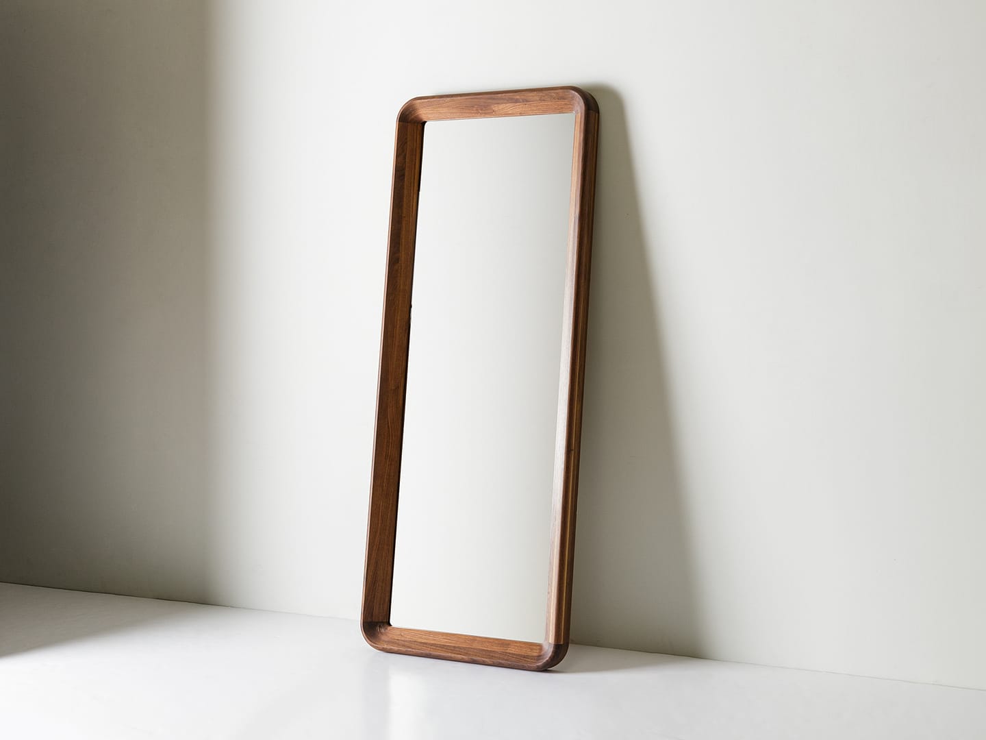 Simple Mirror in walnut tall leaning floor mirror with rounded corner frame leaning at an angle in a white studio corner